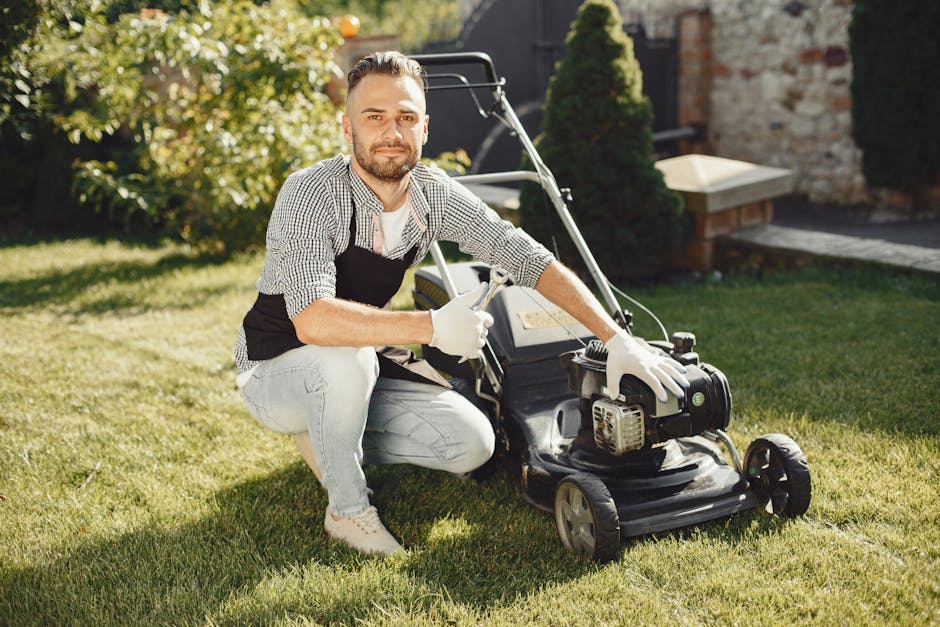 A male gardener with a neatly trimmed beard and short hair, dressed in a checkered shirt, light jeans, a black apron, and white gloves, is kneeling on a lush green lawn, holding a garden blower with a