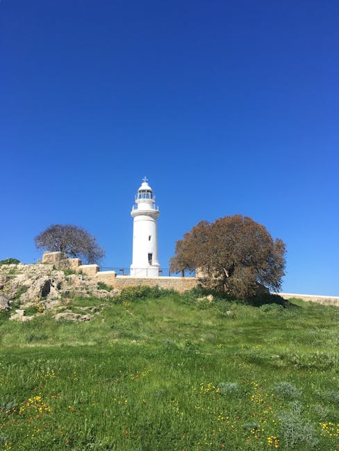A scenic landscape featuring a tall, white lighthouse with a red top situated on a grassy hill. The hill is covered with a mixture of green and yellowish grass, and a narrow, winding dirt path leads u
